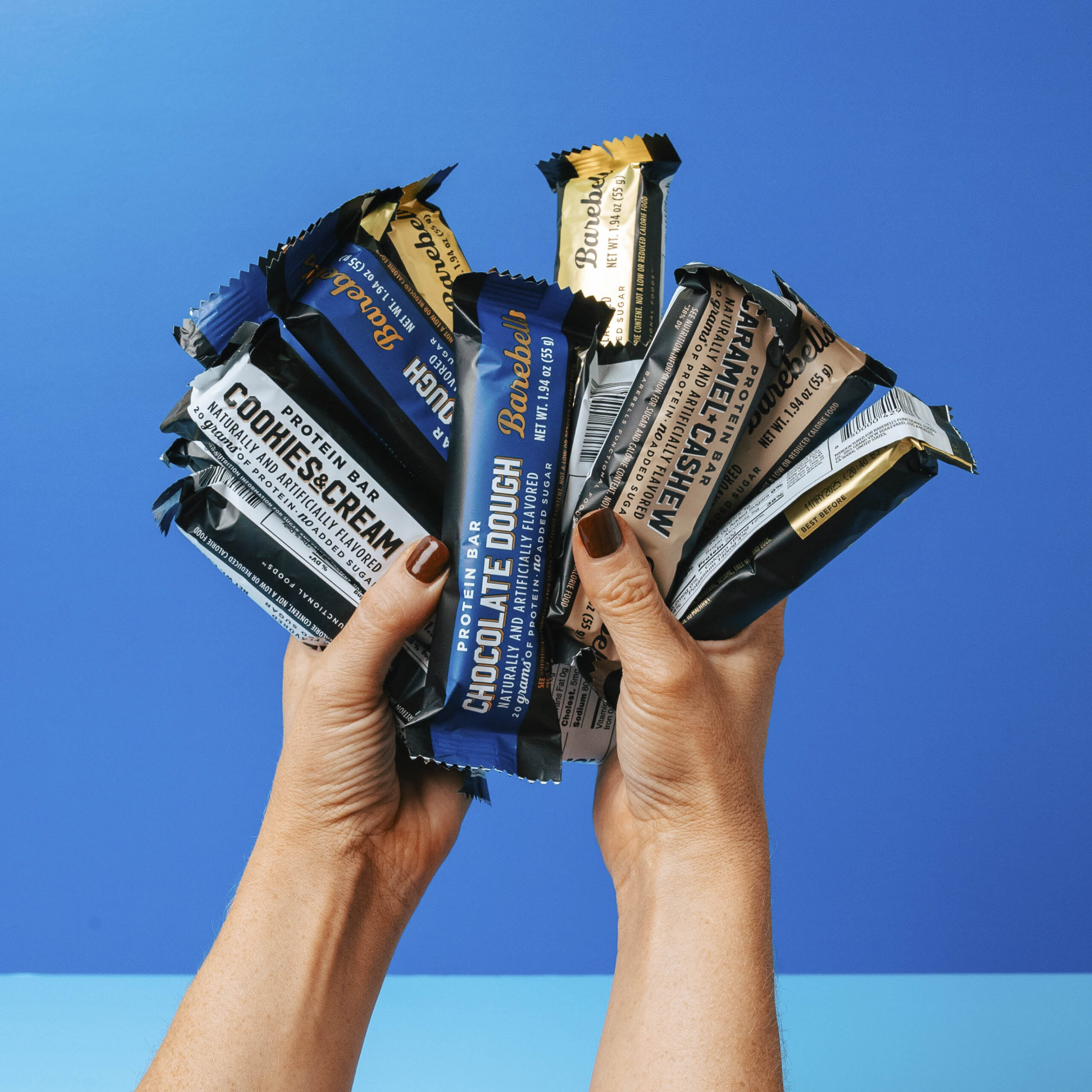 Hands holding multiple protein bars against a blue sky background.